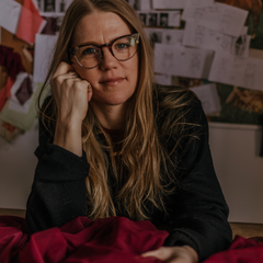 A woman with glasses sitting in front of a camera, with a backdrop featuring various papers and designs, presumably Siobhan Barrett, the owner of Siobhan Barrett Lingerie.